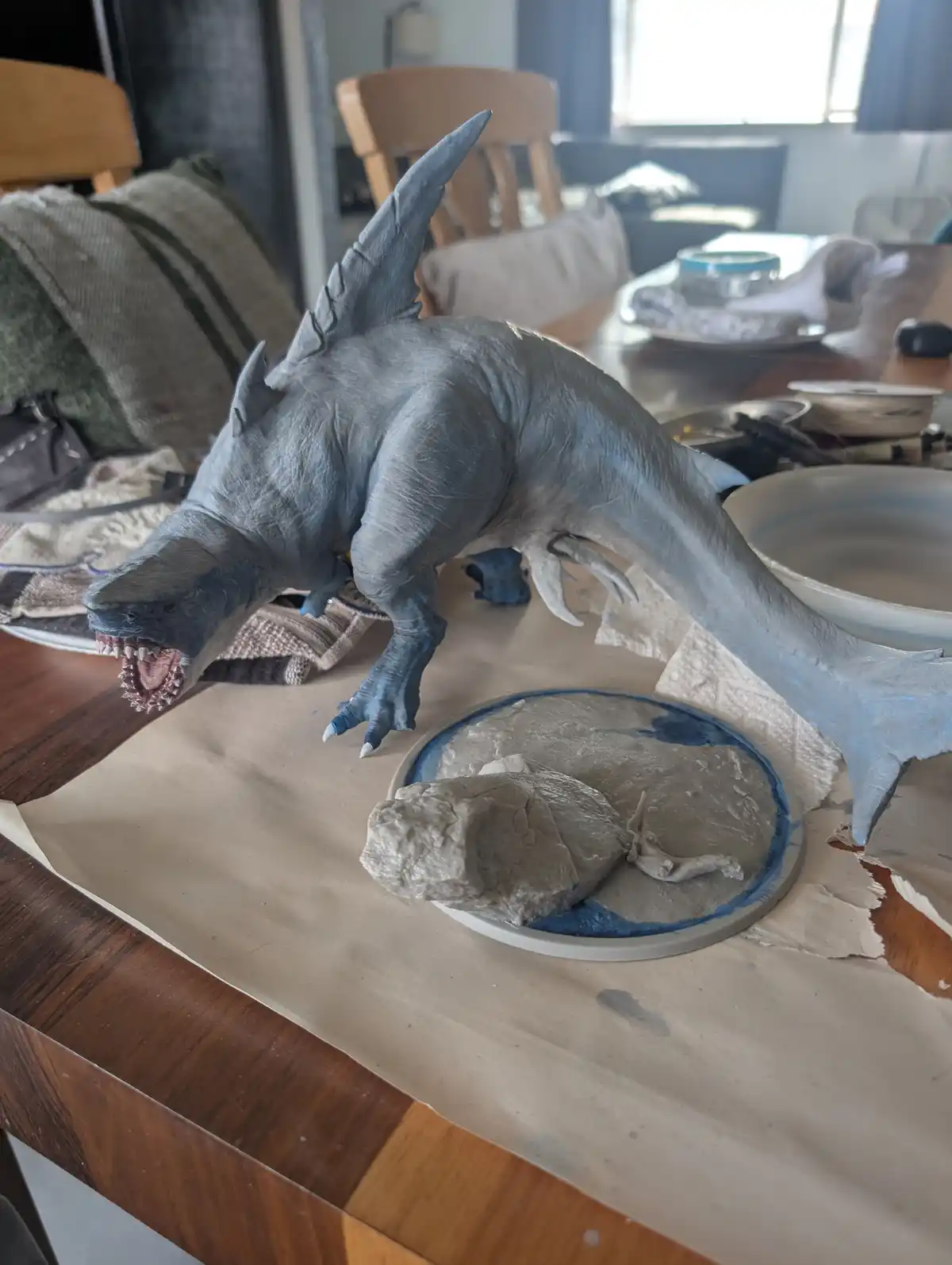 A grey, shark-headed, dinosaur-like creature sculpture with sharp teeth and claws is posed on a wooden table as if attacking…