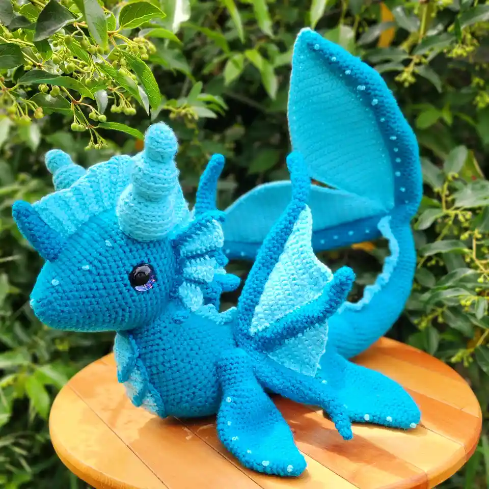 A blue crocheted dragon with fish-like tail and fins sits on a wooden stool outdoors, surrounded by green foliage. The…