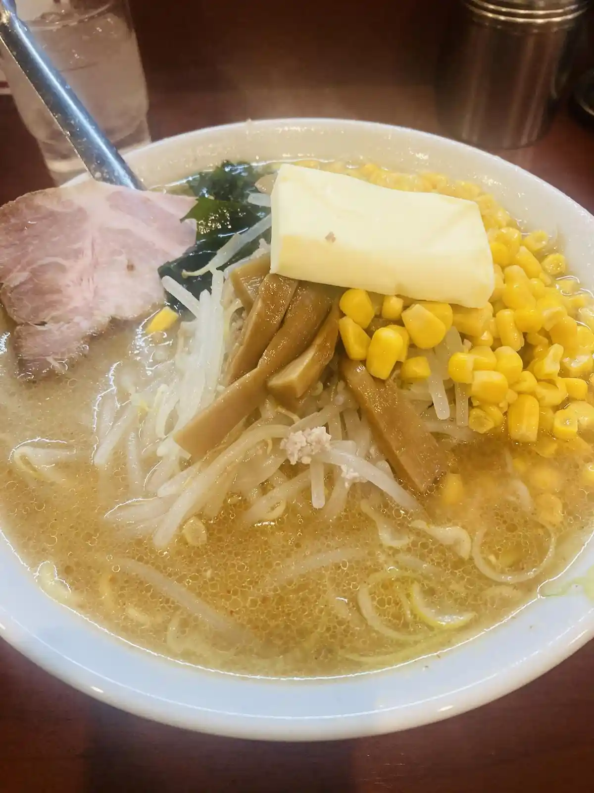 A close-up photo of a steaming bowl of ramen topped with a large, rectangular pat of butter, yellow corn kernels, bean…