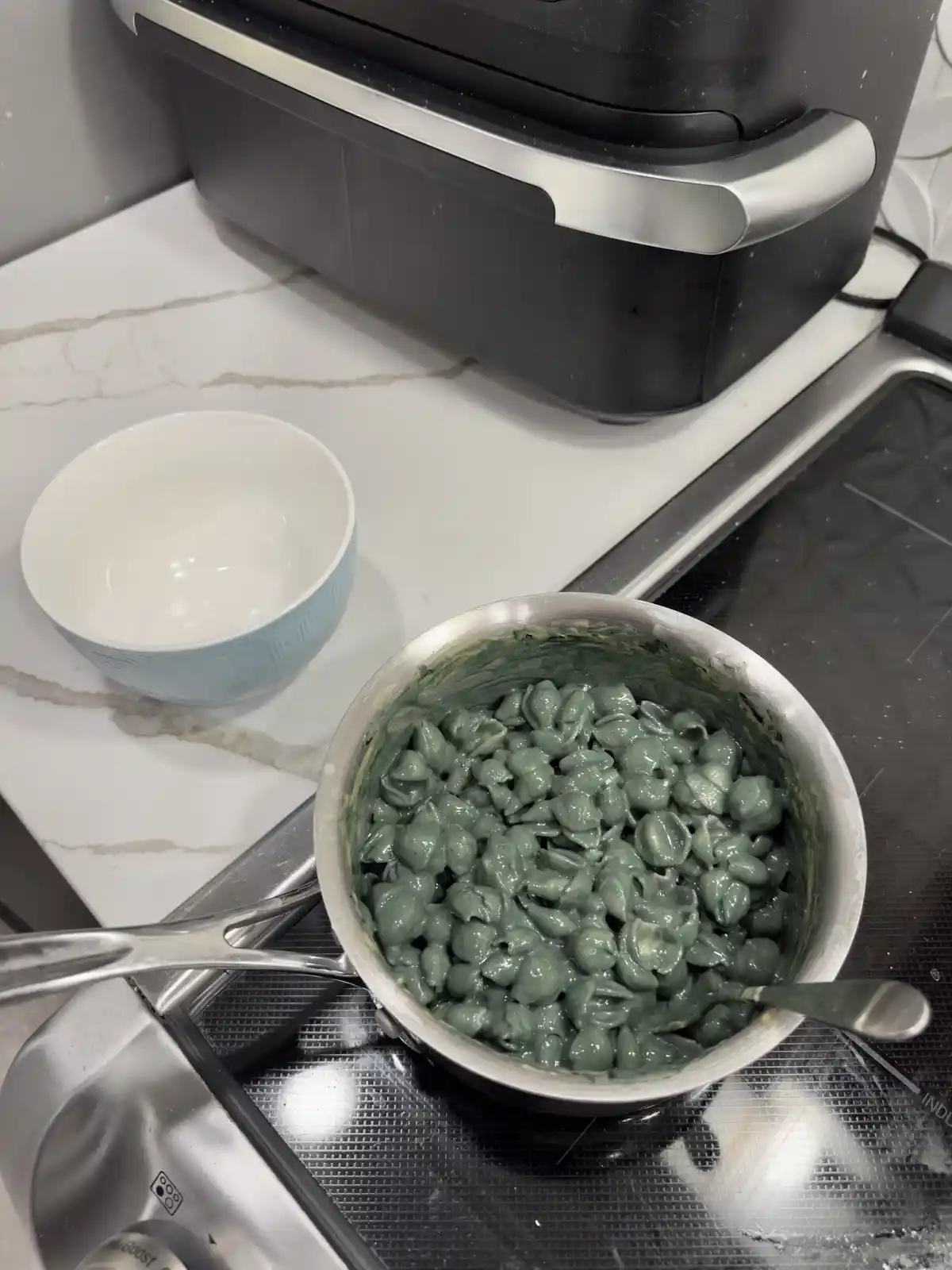 A stainless steel saucepan sits on a stovetop, filled with shell-shaped pasta coated in a thick, green sauce. Beside it on…