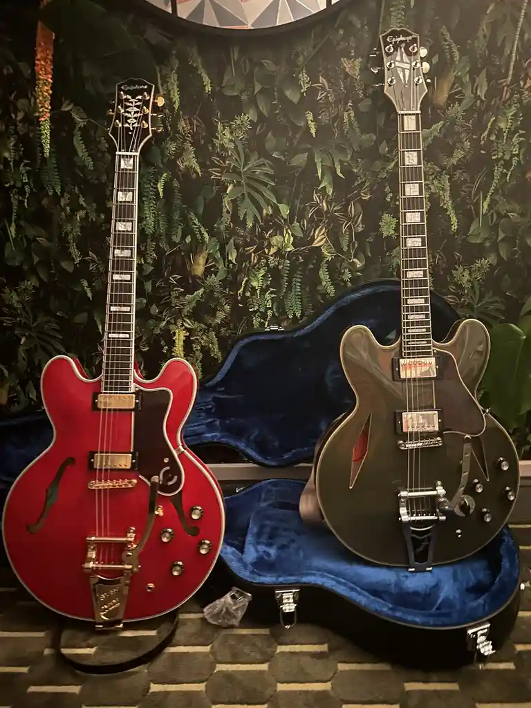 Two Epiphone electric guitars, one red and one dark green, are displayed side-by-side on stands and in open blue cases…