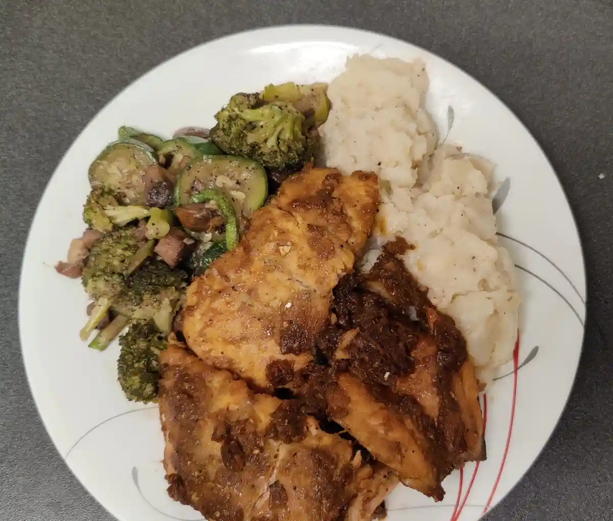 A white plate holds a meal of roasted chicken, mashed potatoes, and mixed vegetables including broccoli and zucchini. The…