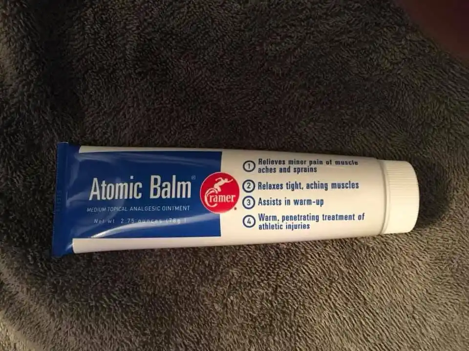 A tube of 'Atomic Balm' topical analgesic ointment by Cramer lies on a dark gray towel. The label lists four numbered…