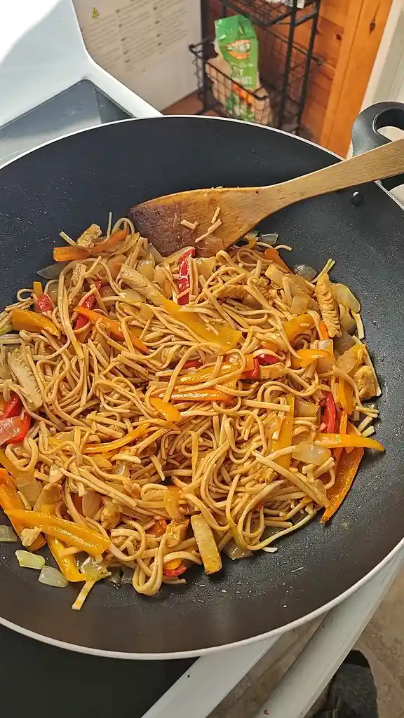 A close-up photo shows a black wok on a stovetop filled with stir-fried noodles, mixed with colorful bell peppers (red and…