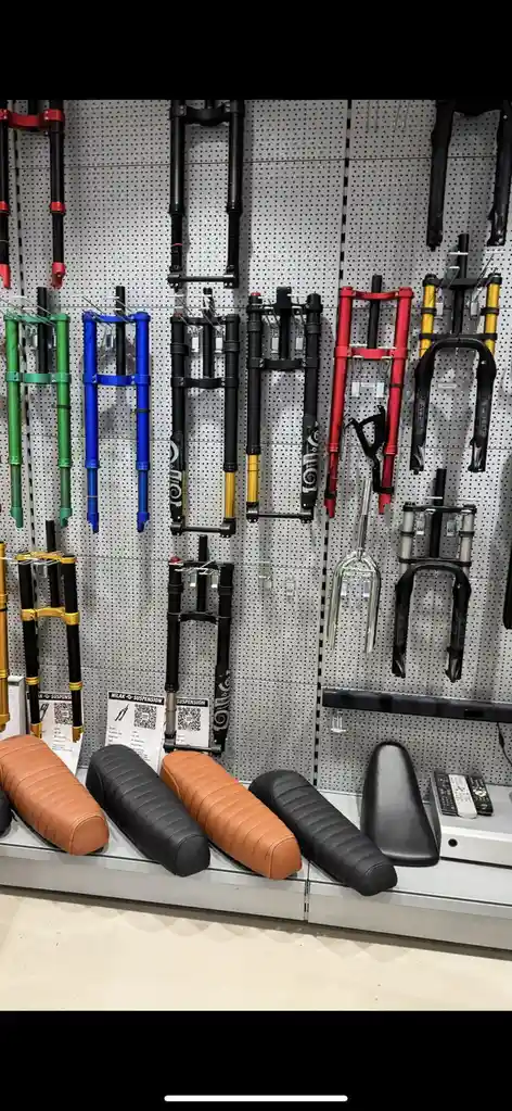 A retail display wall shows numerous colorful bicycle suspension forks hanging vertically on a pegboard. Below them, on a…