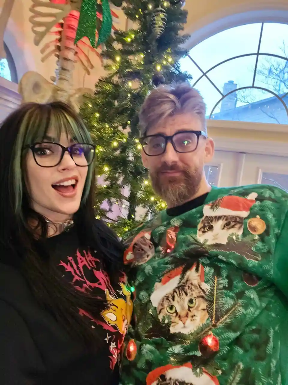 A selfie of a man and a woman in front of a decorated Christmas tree. The man wears a green sweater covered in images of…