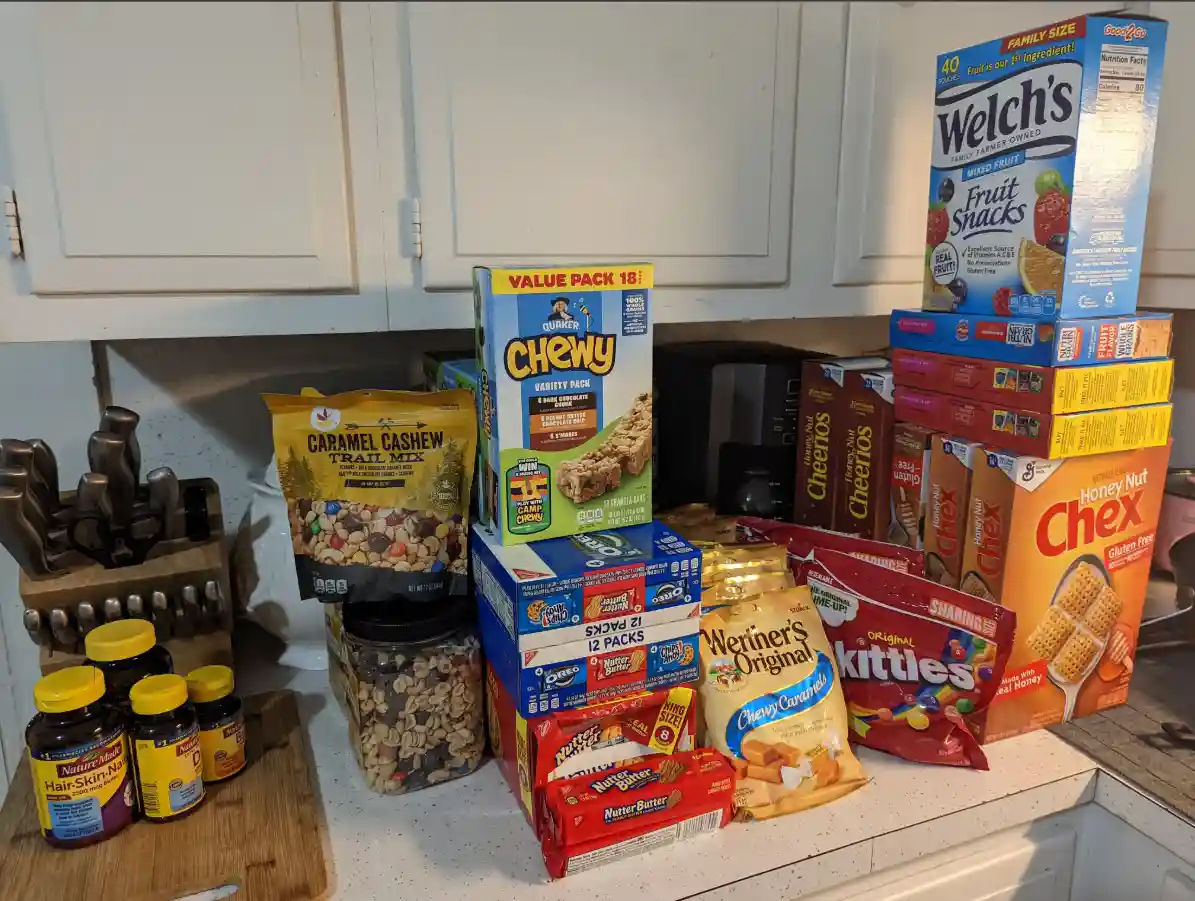 A cluttered kitchen counter is piled high with various snack foods including boxes of Cheerios, Chex, Welch's Fruit Snacks…