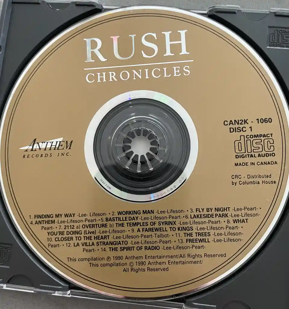 A close-up photo of a Rush 'Chronicles' compilation CD, Disc 1, showing the tracklist and artist credits. The CD is…