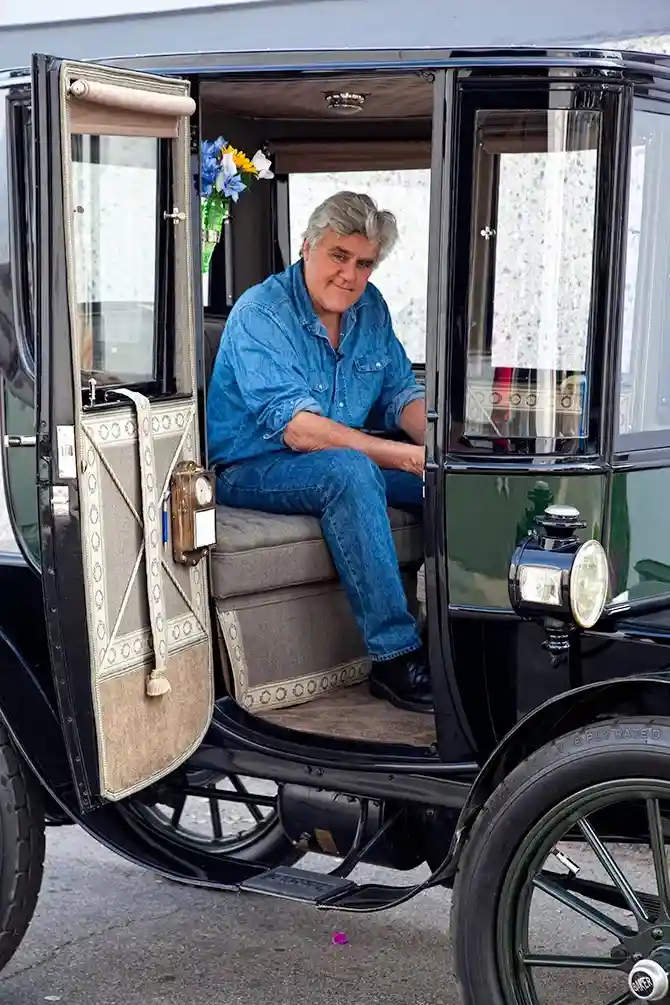 A man with gray hair wearing a blue denim shirt and jeans sits casually in the passenger seat of an old-fashioned black…