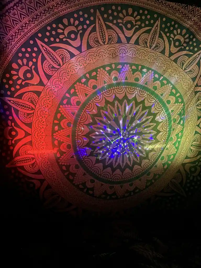 A close-up of a colorful, intricate mandala tapestry hanging on a wall, illuminated by dynamic, multi-colored laser lights…