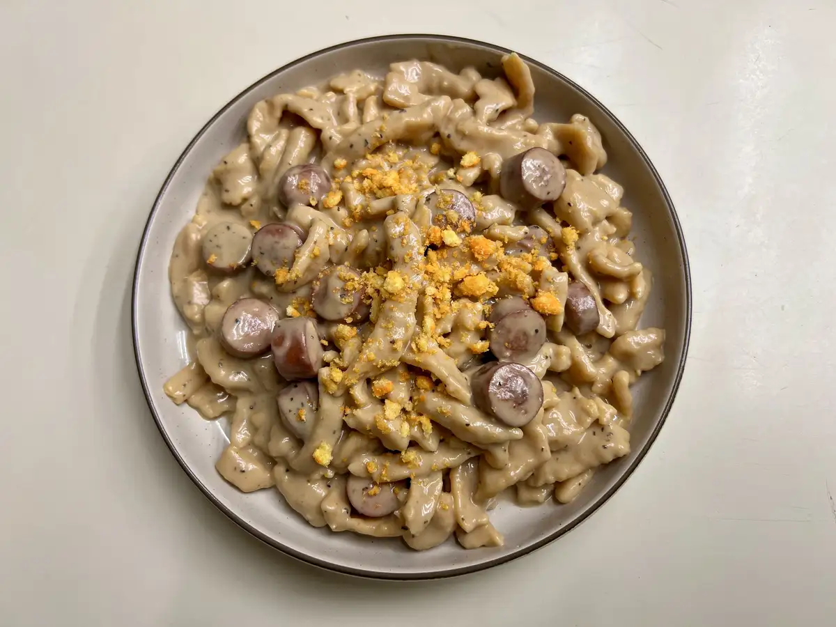 A top-down photo of a plate of creamy pasta mixed with sliced sausages and topped with crushed golden breadcrumbs, with the…