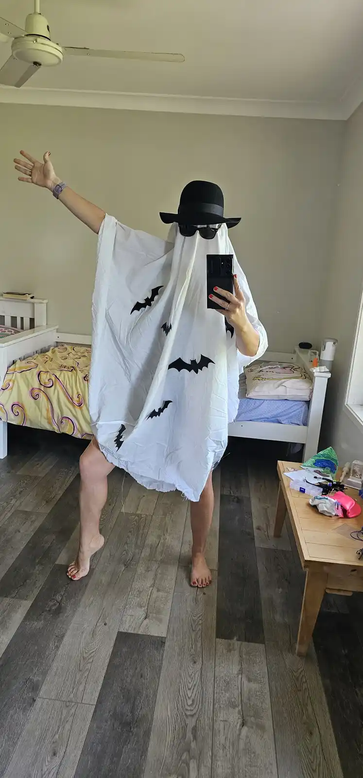 A person in a bedroom takes a mirror selfie wearing a white ghost costume with black bat cutouts, a black bowler hat, and…