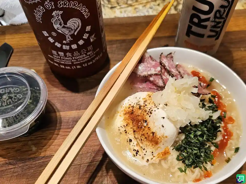 A bowl of ramen with a poached egg, sliced salami, sauerkraut, and green herbs sits on a wooden table with chopsticks…