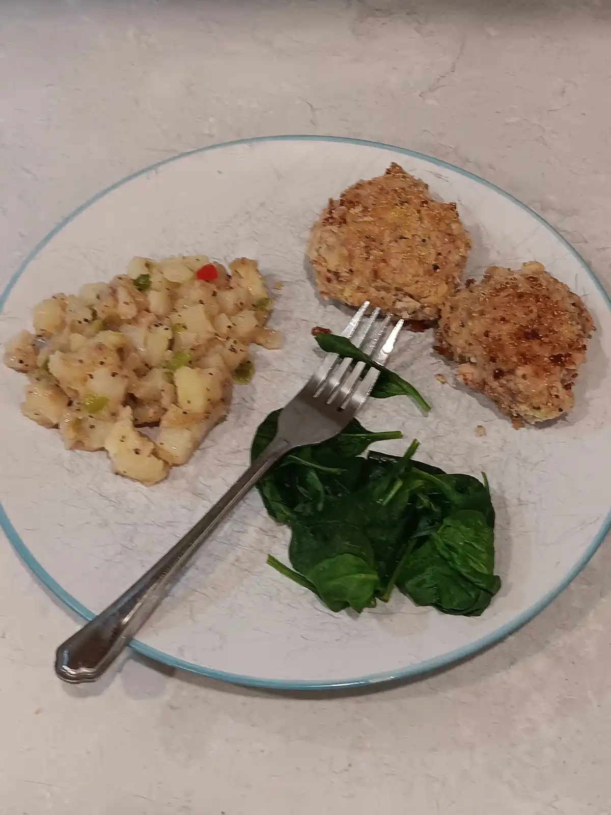 A white plate with a light blue rim holds two golden-brown breaded patties, a serving of diced potatoes with green and red…