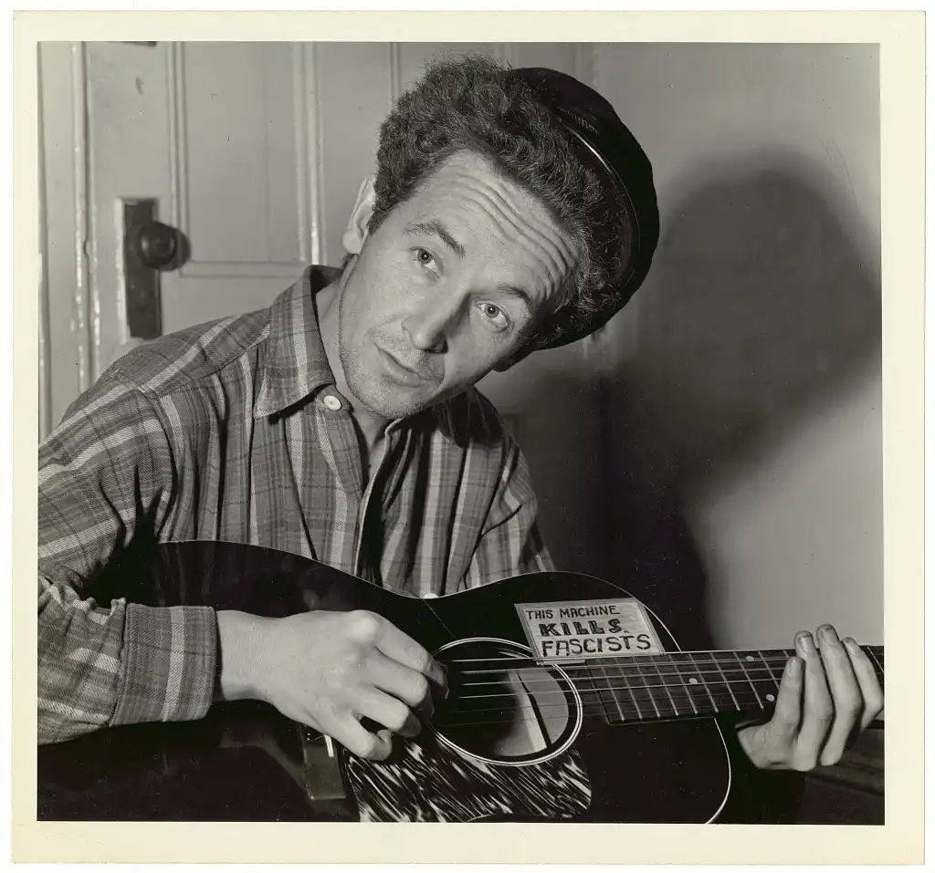 A black-and-white photo of a man in a plaid shirt and cap holding an acoustic guitar. A sticker on the guitar's soundhole…