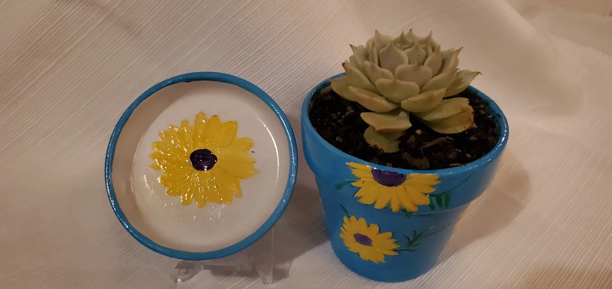 A blue-painted ceramic pot with a painted sunflower design holds a succulent plant, next to its matching blue-rimmed lid…