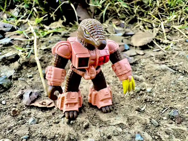 A small, pink and brown toy robot with a reptilian head and yellow claws stands on dirt ground surrounded by grass and…
