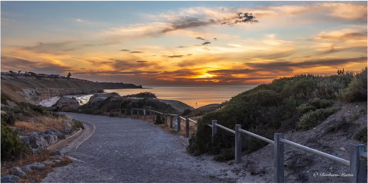 A scenic coastal sunset photo shows a winding gravel path leading toward the ocean, flanked by wooden railings and dunes…