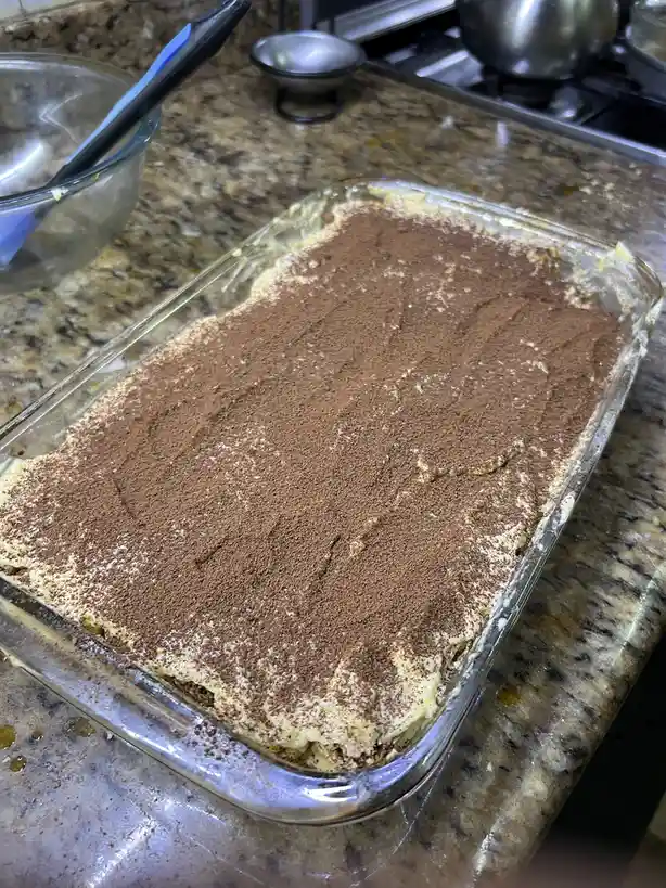 A glass baking dish filled with tiramisu, dusted with cocoa powder, sits on a granite countertop in a kitchen. The dessert…