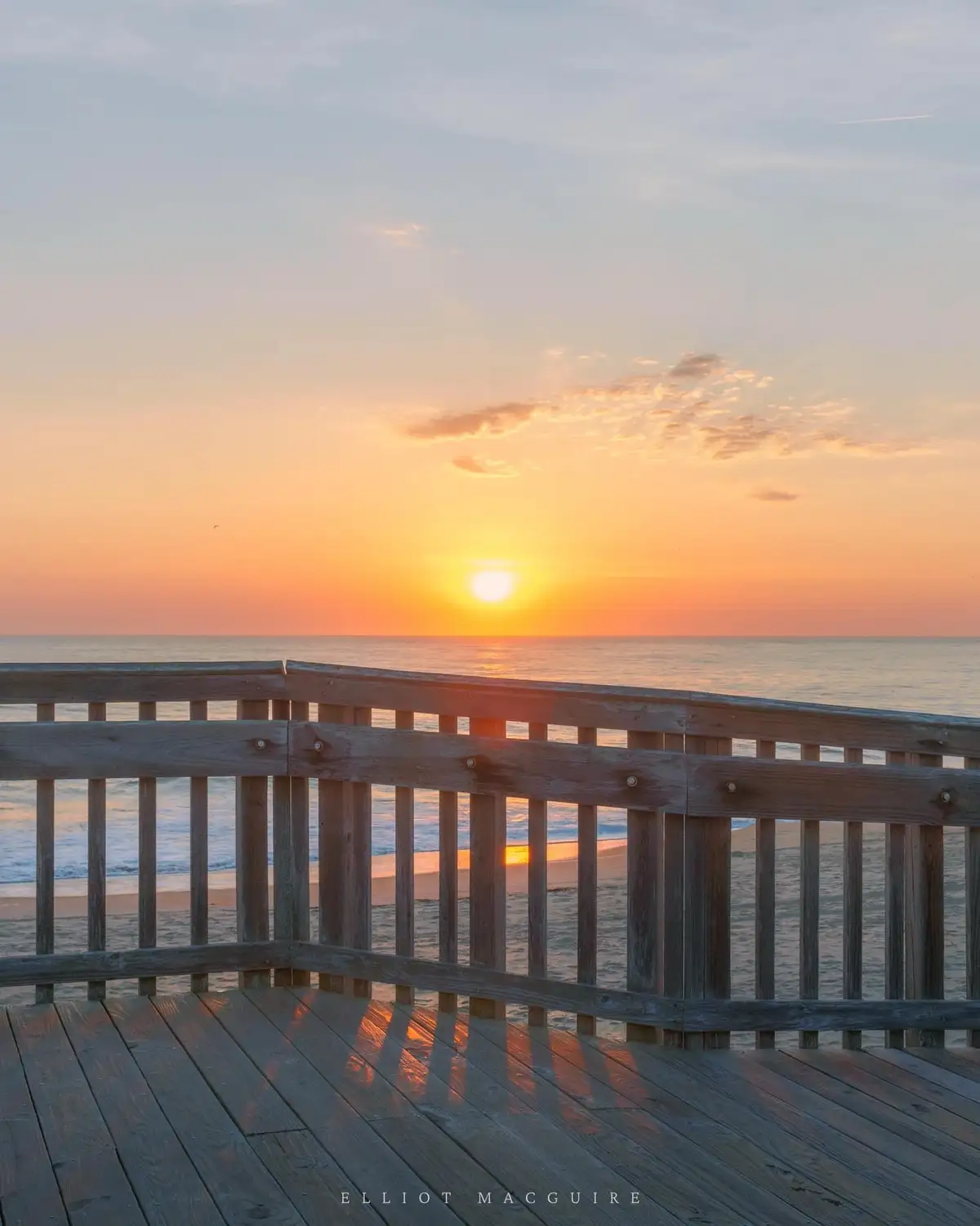 A serene sunset photograph taken from a wooden boardwalk overlooking the ocean, with the sun setting on the horizon and…