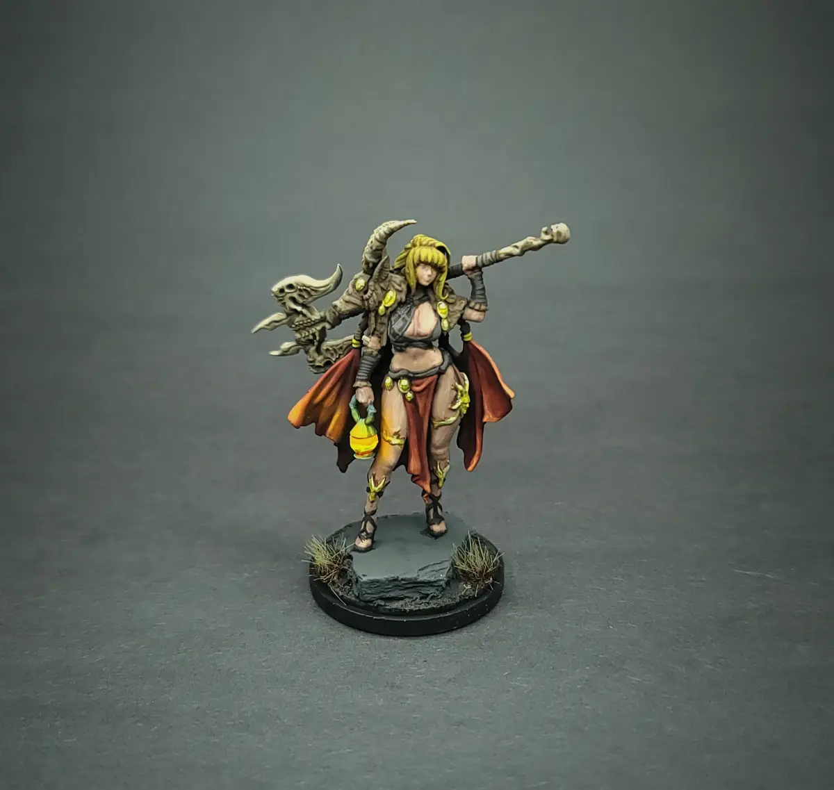 A painted miniature figurine of a female fantasy character with horns, a skull-topped staff, and a red cape, standing on a…