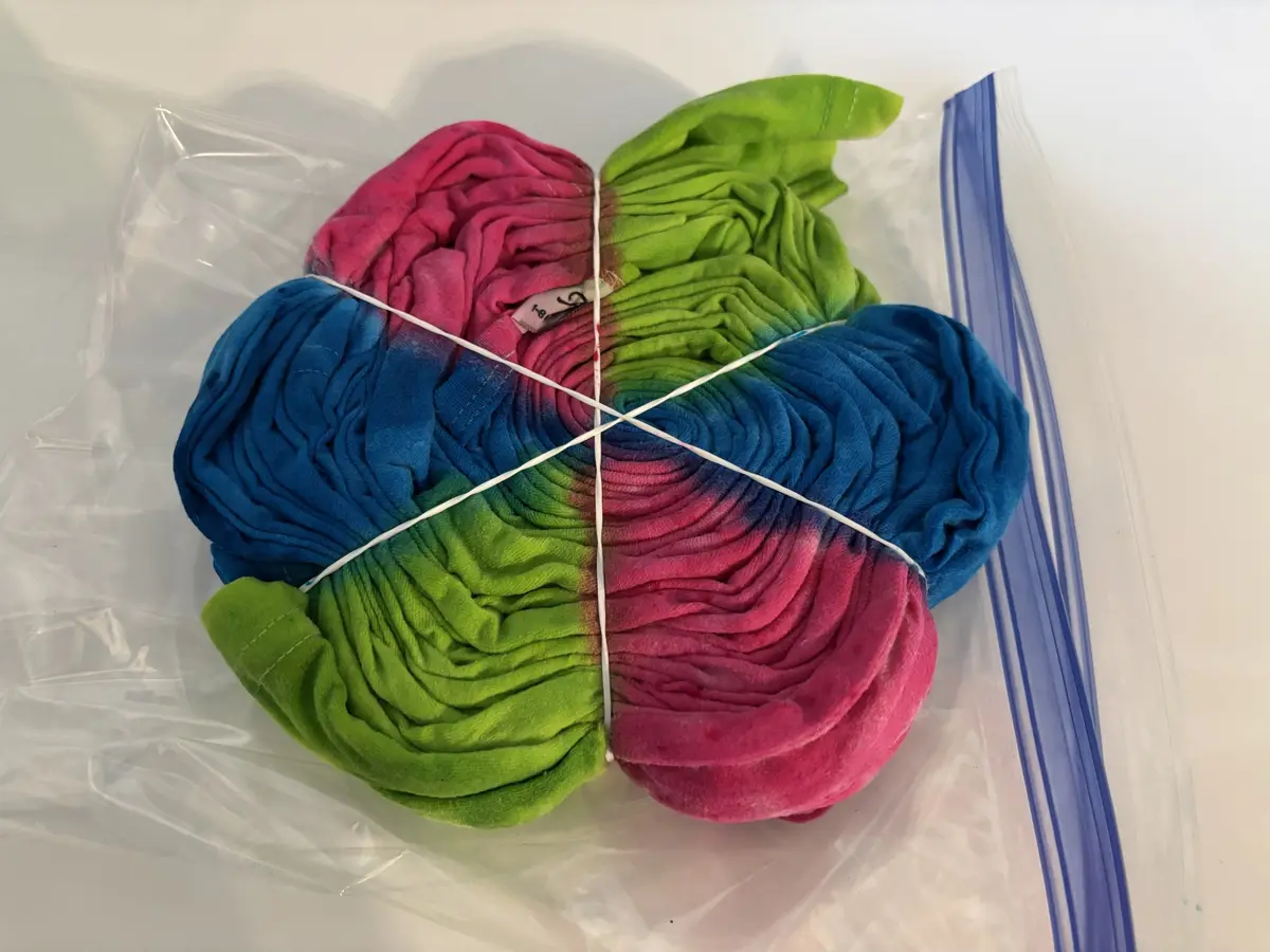 A tie-dyed t-shirt, folded and bound with white rubber bands into a four-petal flower shape, rests inside a clear plastic…