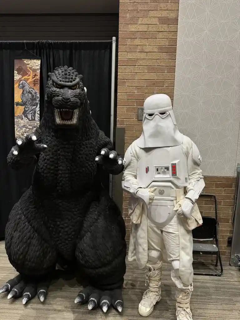 A person in a Godzilla costume stands next to a person in a white stormtrooper costume, both posing for a photo. The…