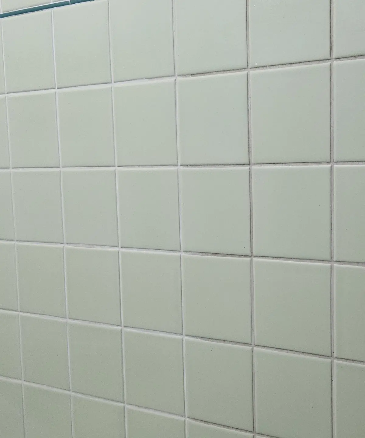 A close-up photo of a wall covered in light green square tiles with white grout lines, showing a repetitive, mundane…