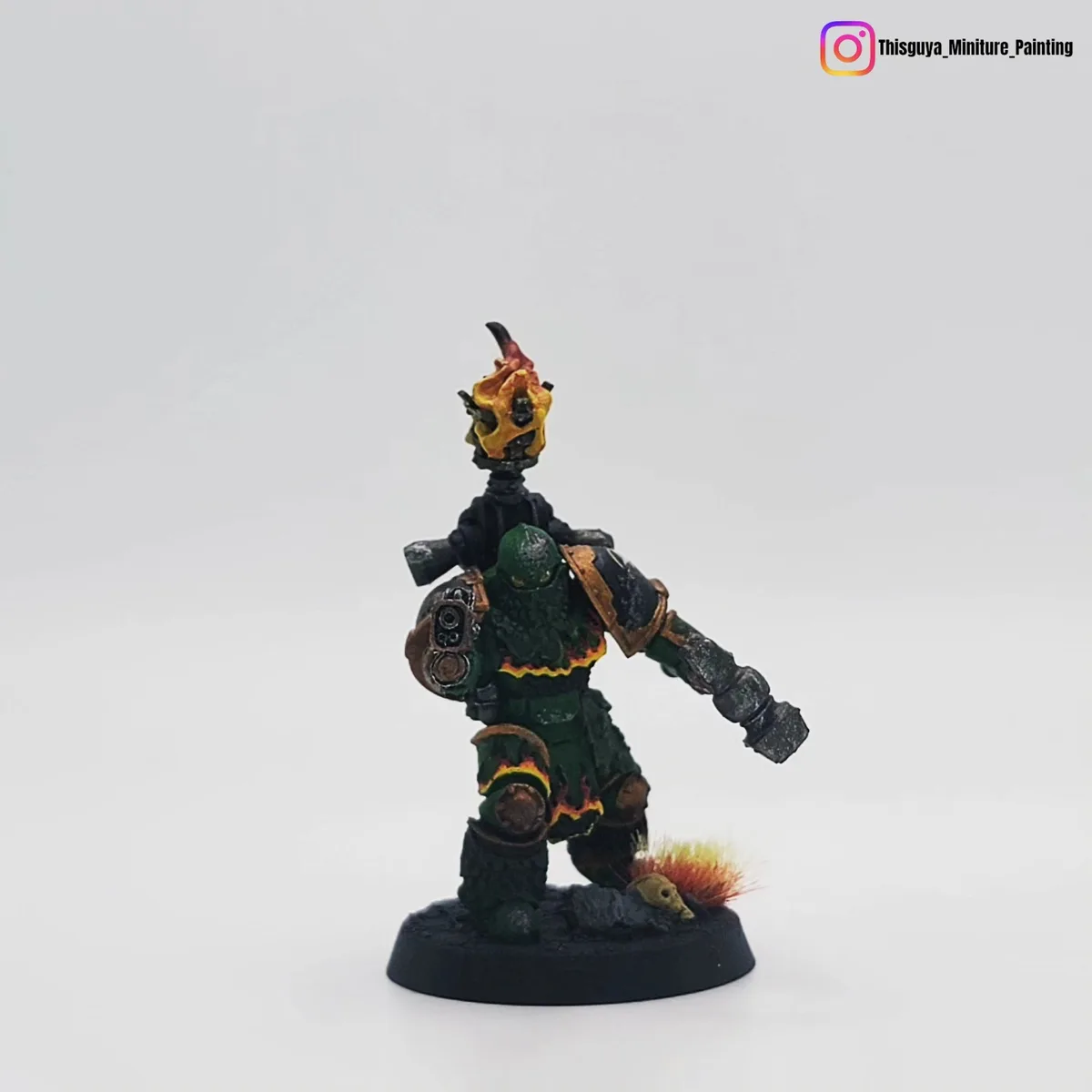 A painted miniature figure of a green-armored warrior with a flaming skull mounted on his shoulder, standing on a black…