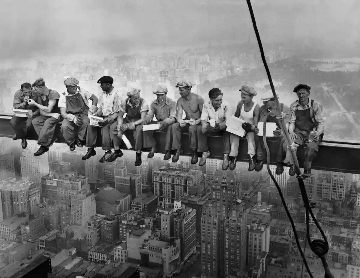 A black-and-white historical photo shows ten construction workers casually sitting on a steel beam high above a dense…