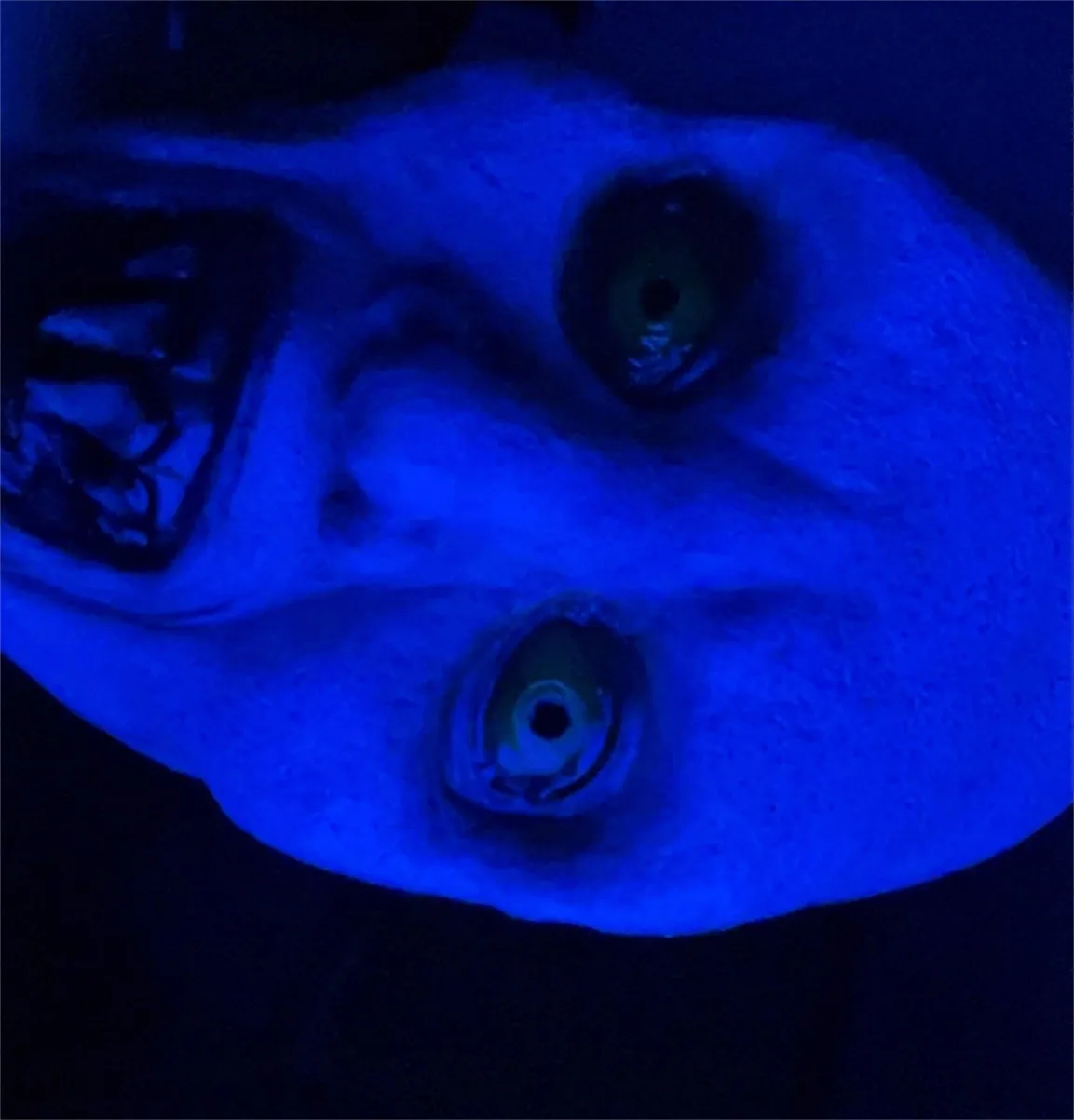 A close-up of a person's face under blue light, showing wide, glowing eyes and an open mouth, conveying a startled or…