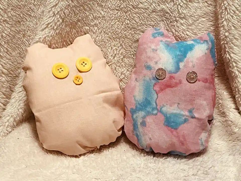 Two handmade cat-shaped pillows sit side by side on a fluffy beige blanket. The left pillow is plain beige fabric with three…