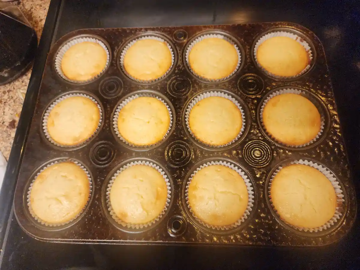 A close-up photo shows a dozen freshly baked, golden-brown cupcakes in a dark, well-used muffin tin, each nestled in a white…