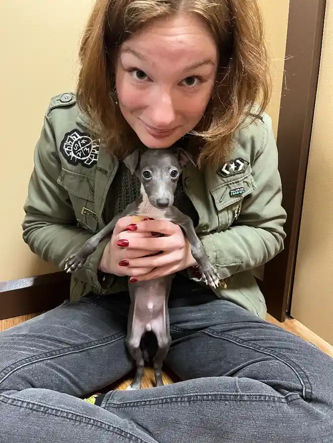 A woman in a green military-style jacket and jeans is sitting on the floor holding a small, slender grey puppy. She is…