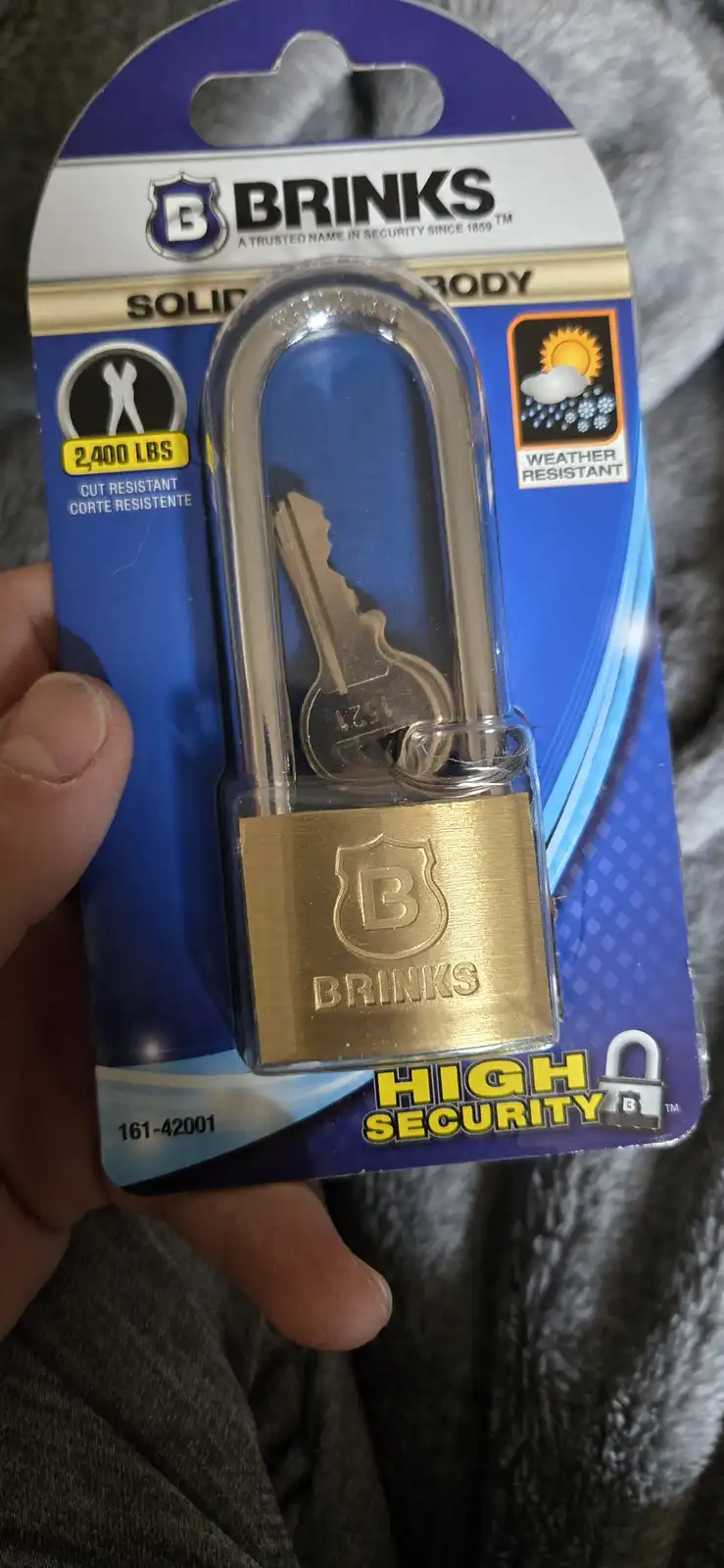 A hand holds a Brinks brand high-security padlock in its blue and gold retail packaging. The lock is advertised as '2,400…