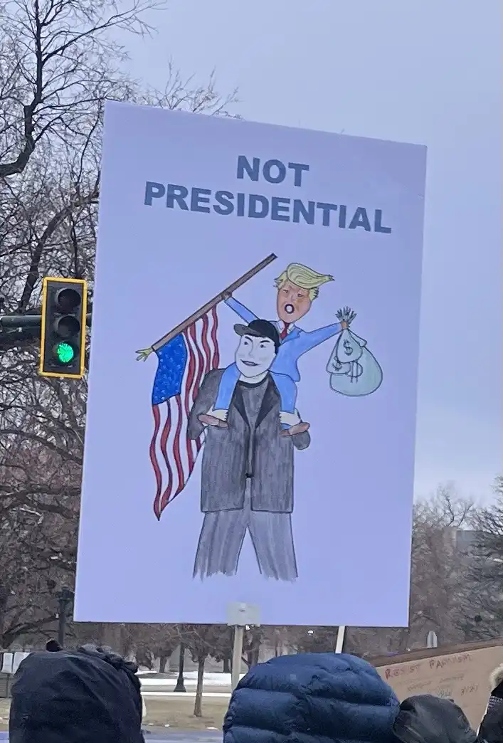A protest sign shows a cartoon of Donald Trump riding piggyback on a man in a suit, holding an American flag and a money…