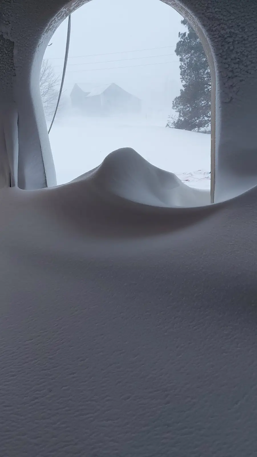 A view from inside a snow-covered structure looking out through a circular opening, where a thick, sculpted mound of snow…
