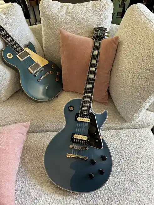 Two blue electric guitars, resembling Gibson Les Paul models, are resting on a light-colored, textured couch cushioned with…