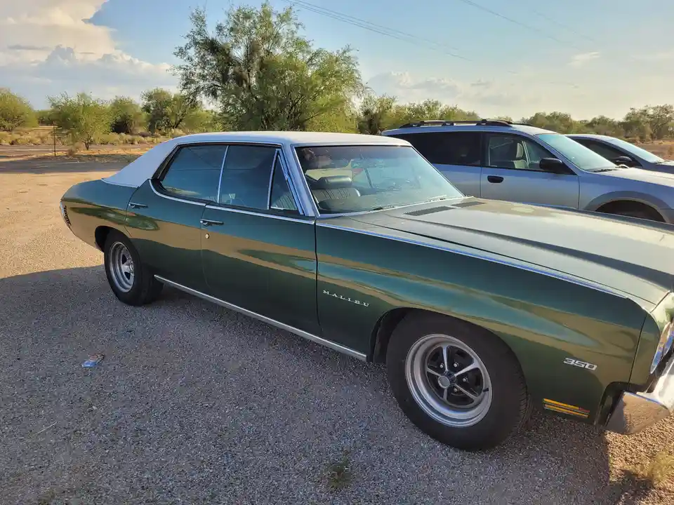 A green 1960s Chevrolet Malibu sedan with a white roof is parked on gravel, featuring 'MALIBU' and '350' badges. Behind it…
