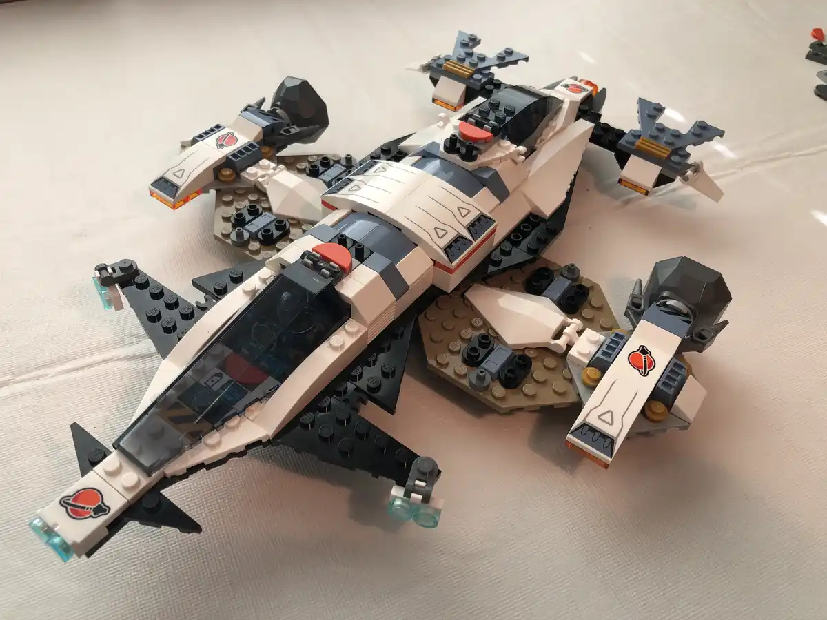 A white, black, and gray LEGO Star Wars fighter jet with orange and translucent blue details rests on a light-colored fabric…