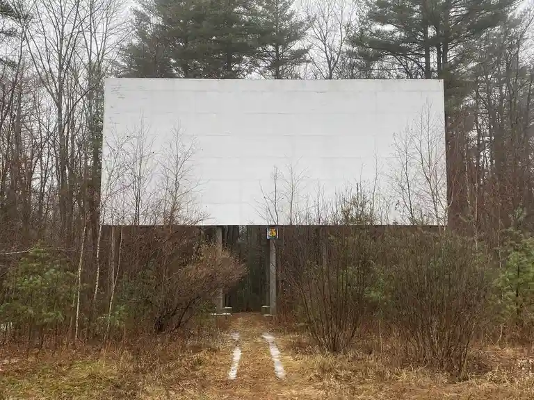 A large, blank white outdoor movie screen stands in a wooded area, surrounded by bare trees and shrubs, with a muddy dirt…