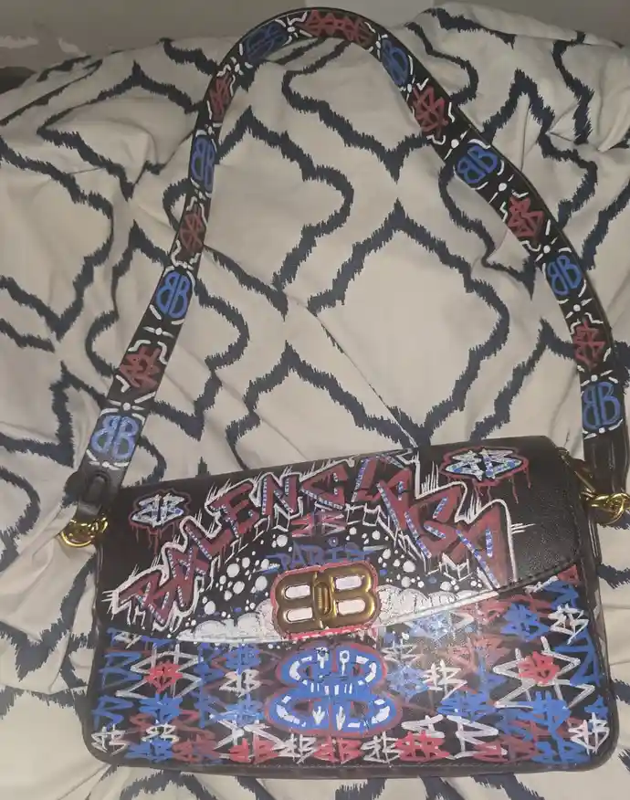 A black Balenciaga handbag with a graffiti-style design featuring red, blue, and white spray-painted lettering and logos…