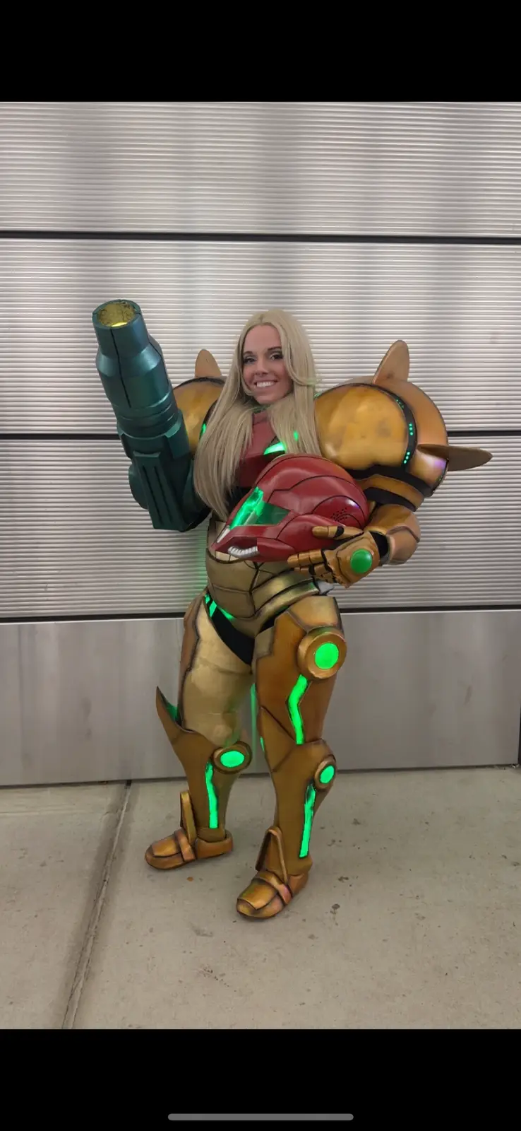 A person with long blonde hair is posing in a detailed, full-body Samus Aran cosplay from Metroid, featuring gold armor with…