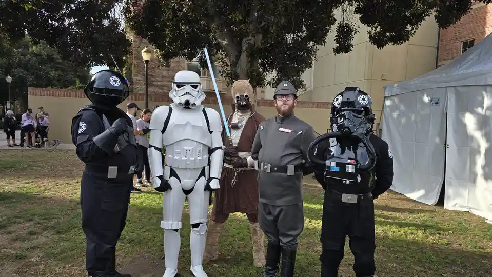 Five people in Star Wars costumes stand outdoors under a tree: a black-armored Imperial officer, a white Stormtrooper…