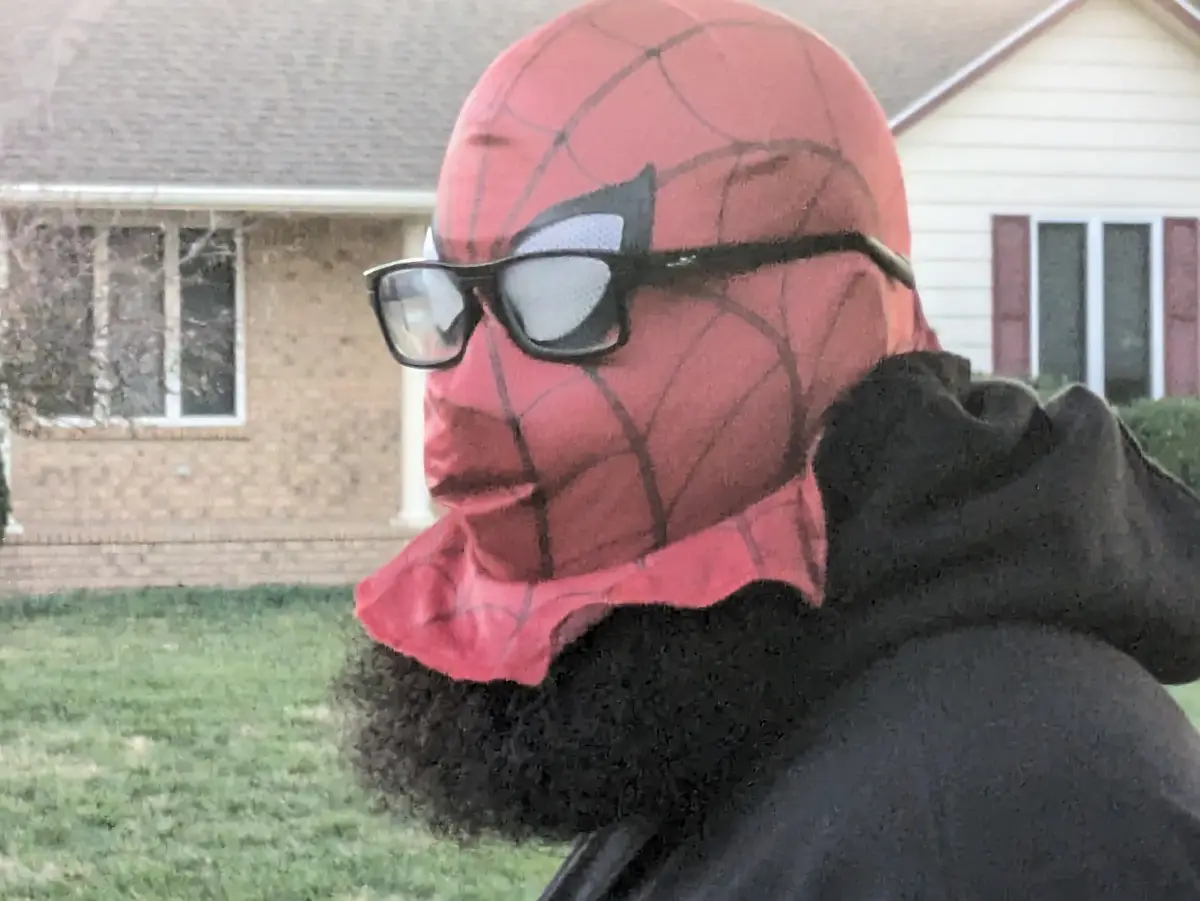A person wearing a red Spider-Man mask with black webbing and black-rimmed glasses, standing outdoors in front of a house…
