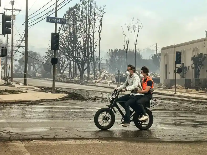 Two men wearing face masks ride an electric bicycle down a wet street in a burned-out area, with a 'Sunset Bl' street sign…