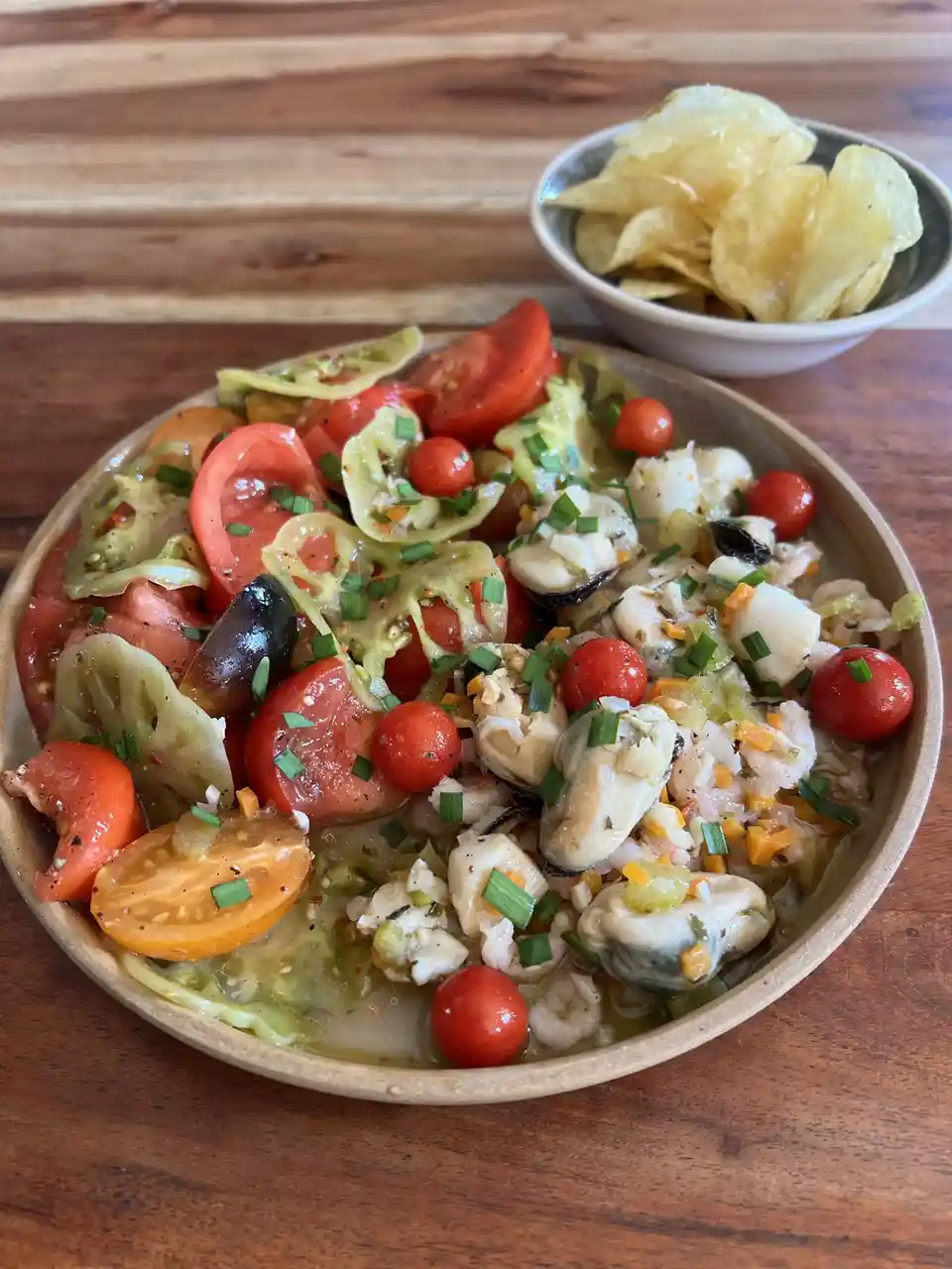 A wooden bowl filled with a colorful seafood salad featuring oysters, shrimp, cherry tomatoes, and sliced heirloom tomatoes…
