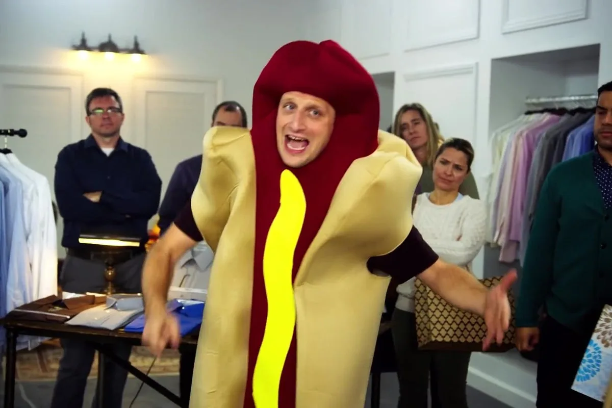 A man in a full-body hot dog costume with a yellow mustard stripe and red bun is posing dramatically with arms outstretched…