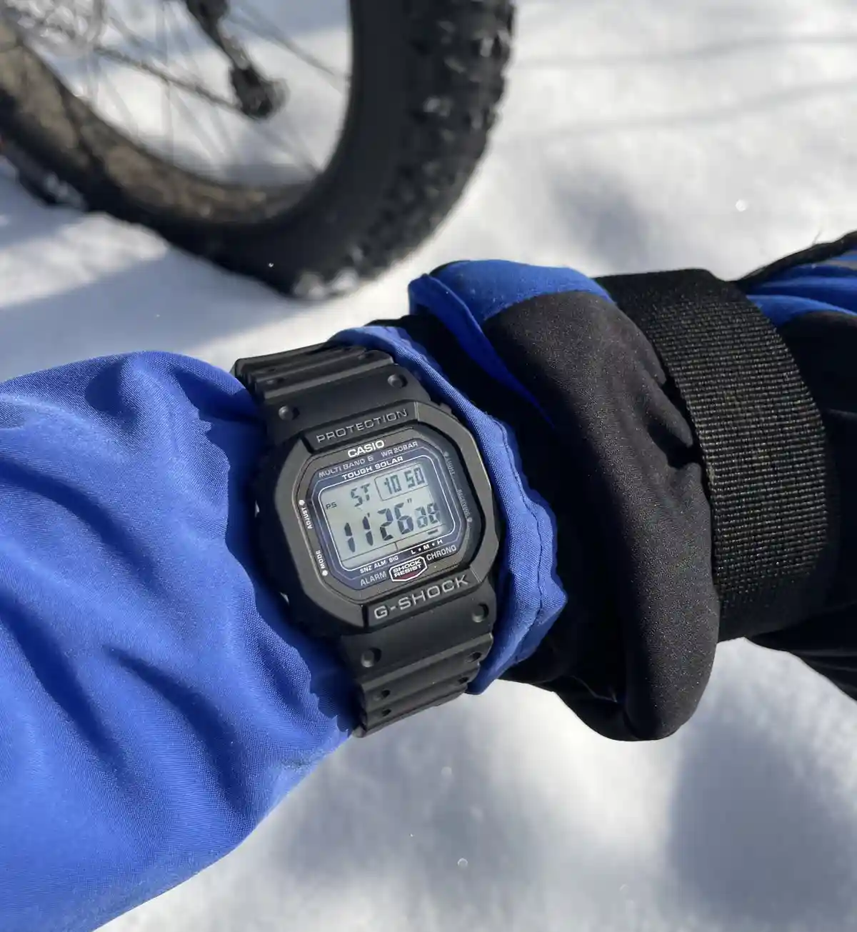 A close-up of a person's gloved hand wearing a black Casio G-Shock watch, displaying the time 11:26:00, set against a snowy…