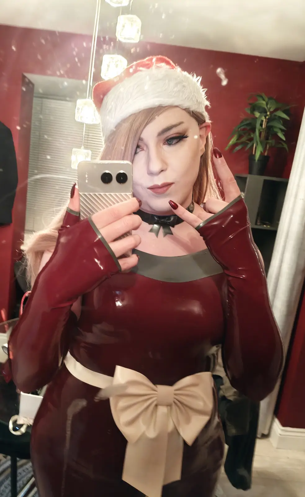 A person in a shiny red latex outfit with a large beige bow at the waist and a Santa hat takes a mirror selfie with a…